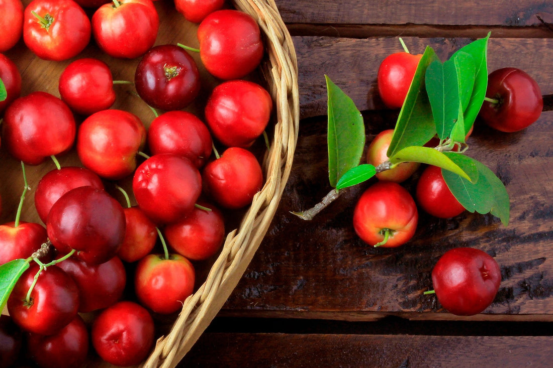 A woven basket of ripe red acerola cherries on a rustic dark wood table, with a few cherries and green leaves beside it