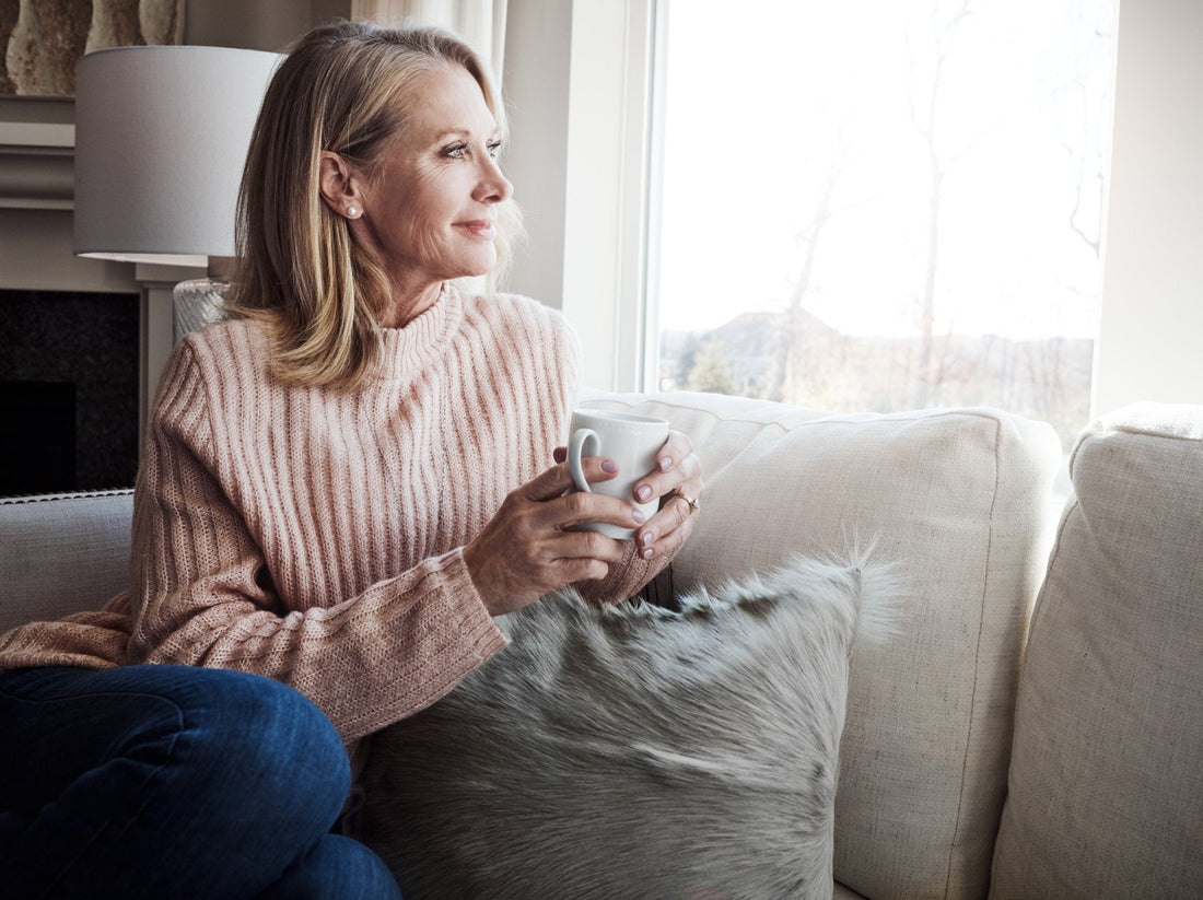 A woman in her late fifties sitting on a sofa by a bright window in a soft pink sweater, holding a mug of tea and looking out at the winter trees