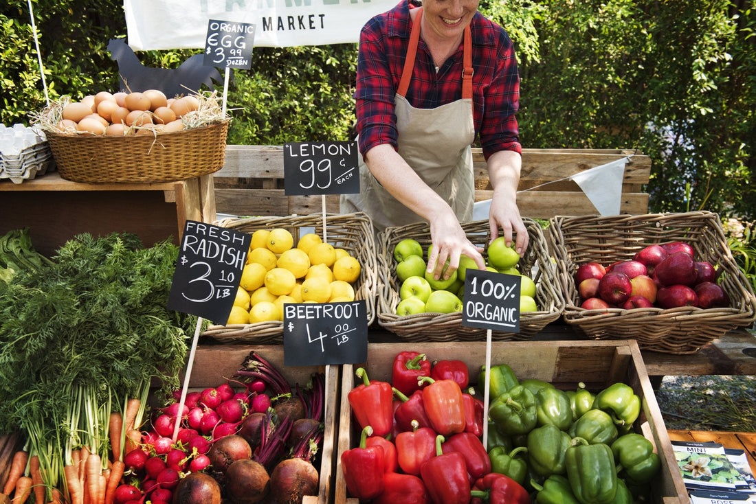A market vendor arranging baskets of apples at a farmers market stall surrounded by handwritten chalkboard price signs, bell peppers, radishes, and beets