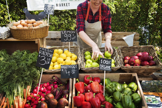 A market vendor arranging baskets of apples at a farmers market stall surrounded by handwritten chalkboard price signs, bell peppers, radishes, and beets
