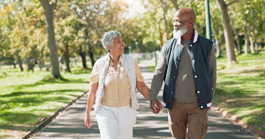 A smiling couple in their sixties holding hands on a tree-lined park path on a sunny day