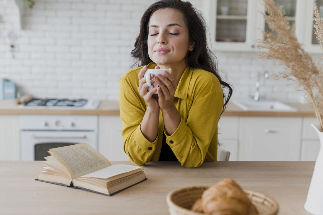 A woman in her forties sitting at a kitchen table in morning light with both hands wrapped around a mug of tea, eyes closed, shoulders relaxed